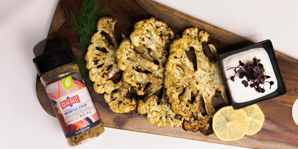 Roasted cauliflower steaks seasoned with herbs and spices are presented on a wooden cutting board alongside a small bowl of white dipping sauce and lemon slices. 
