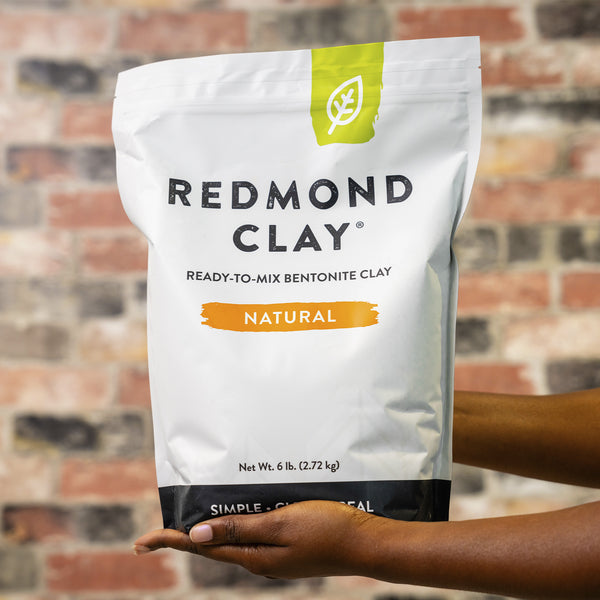 Hands hold a six-pound bag of Redmond Clay, a natural, ready-to-mix bentonite clay product.  
