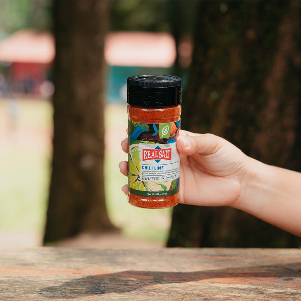 Hand holds a jar of Real Salt Chili Lime seasoning over a picnic table outdoors. 
