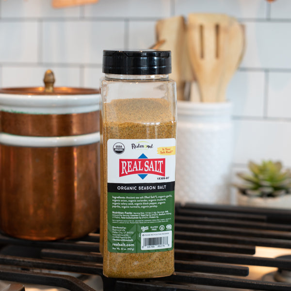 A jar of Redmond Real Salt Organic Season Salt sits on a stovetop, showcasing its blend of ancient sea salt and organic spices. 
