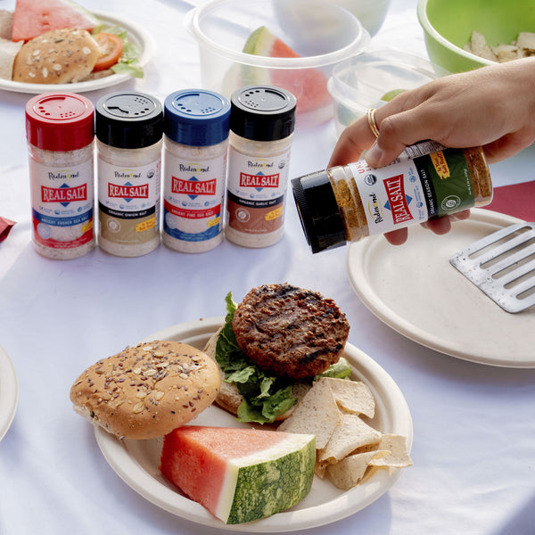 Hand shakes Redmond Real Salt Organic Seasoning Salt over a juicy burger on a plate with watermelon and chips. 

