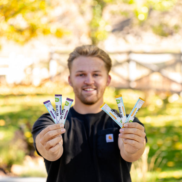 A smiling man holds up three different flavors of Re-Lyte electrolyte powder sticks. 
