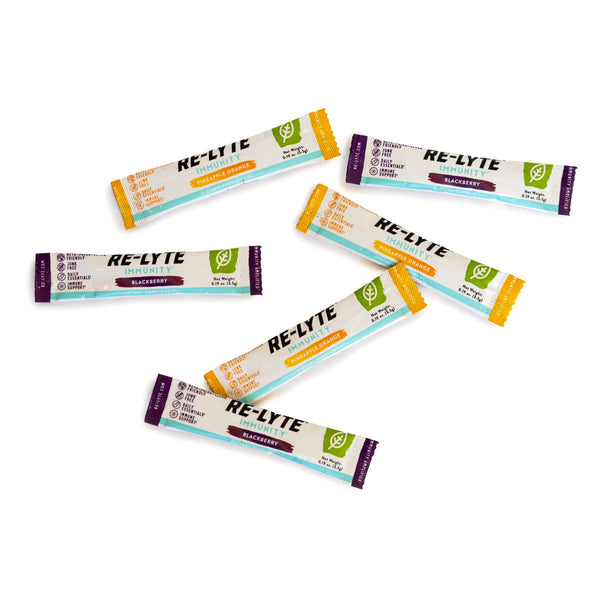 Seven single-serving packets of Re-Lyte Immunity electrolyte powder in Blackberry and Pineapple Orange flavors lay scattered on a white surface. 
