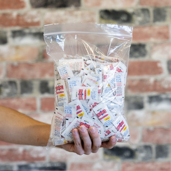 Hand holds a clear plastic bag filled with numerous individual packets of Real Salt brand sea salt. 
