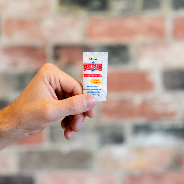 Hand holds a single-serving packet of Redmond Real Salt against a blurred brick background. 
