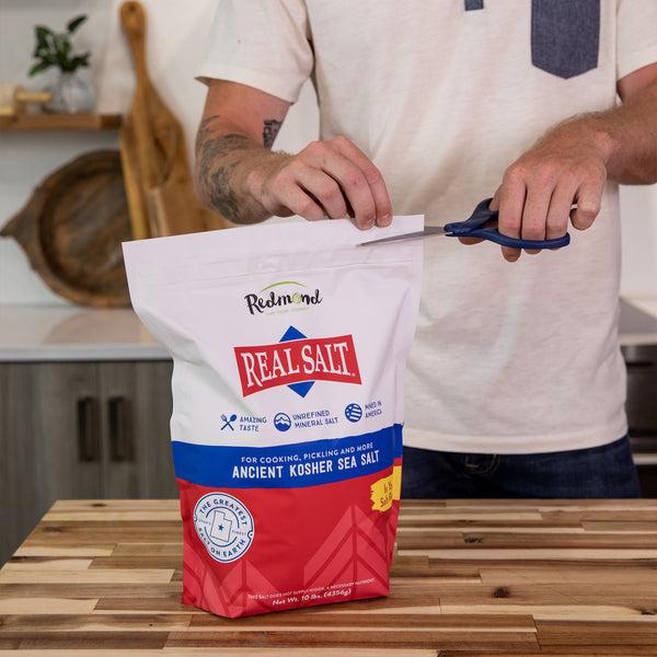 Man opens a 10lb bag of Redmond Real Salt Ancient Kosher Sea Salt with scissors. 
