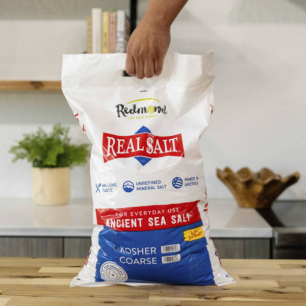 Hand holding a large bag of Redmond Real Salt Ancient Sea Salt, Kosher Coarse. 
