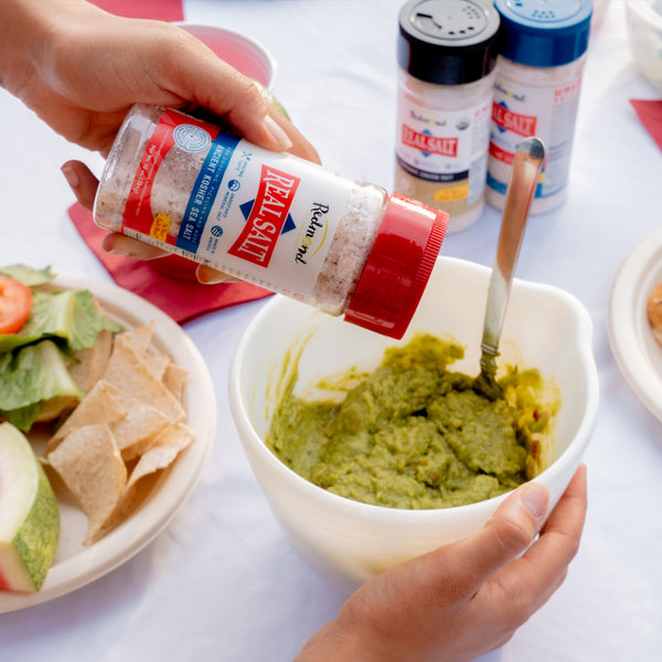 A hand sprinkles Redmond Real Salt Ancient Kosher Sea Salt into a bowl of fresh guacamole. 
