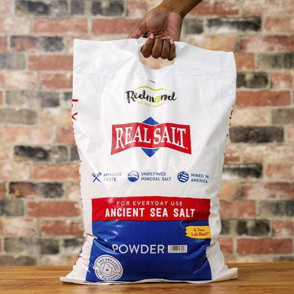 Hand holds a large bag of Redmond Real Salt Ancient Sea Salt Powder. 
