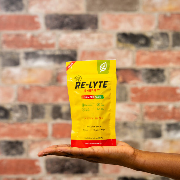 Hand holds a bright yellow Re-Lyte Energy sample pack against a brick wall. 
