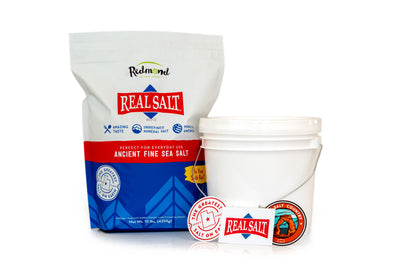 A bag of Redmond Real Salt Ancient Fine Sea Salt stands beside an empty white bucket, showcasing the product ready for use. 
